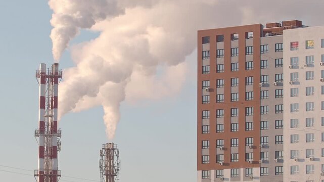Thermal power plant pipes with vapor rising to the sky near the new residential building. Vapor rising in front of the blue sunny sky.