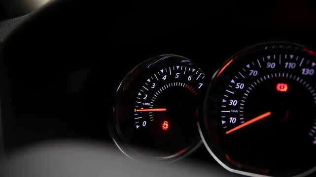 Illuminated car dashboard gauges at night. Tachometer shows engine idling. Open door warning active. Speedometer zero km/h. Parking brake engaged. Vehicle stationary. Operational detail.