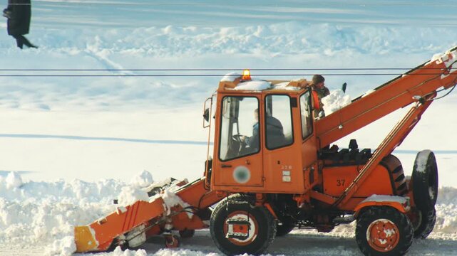 View of an orange snow loader working to clear large amounts of snow from an open area during winter. One person operates machine from cab, another stands on side, supervising operation. Bright day