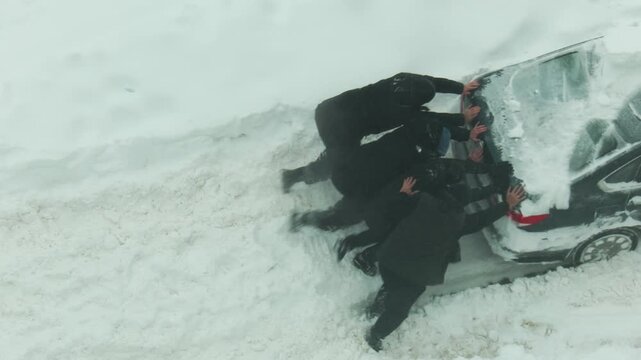 Aerial view of four individuals pushing black car deeply embedded in heavy snowdrift on winter road, demonstrating struggle and teamwork to clear vehicle during extreme weather event