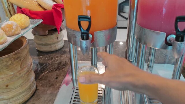 Close-up of a hand pouring fresh orange juice from a beverage dispenser into a glass at a hotel breakfast buffet. Bakery bread and pastries are visible in the background. Concept of self-service dinin