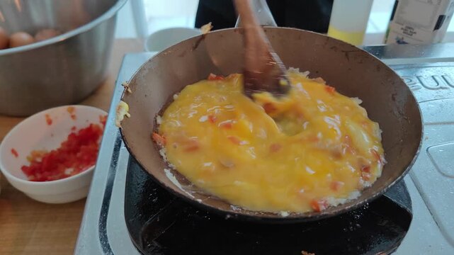 Close-up of pouring beaten egg into a frying pan while cooking scrambled eggs on a stove. A wooden spatula is used to stir the eggs during the cooking process. Concept of breakfast preparation, home c