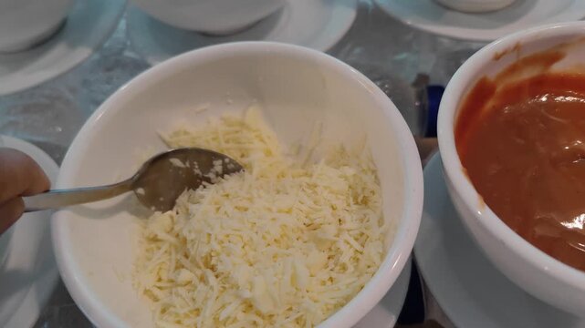 Close-up shot of freshly grated cheese held on a spoon above a white bowl filled with shredded cheese. A bowl of tomato sauce is visible beside it, creating a cooking and food preparation concept. Per