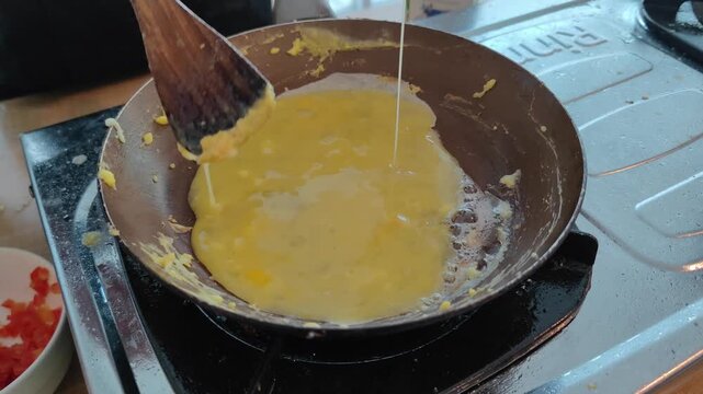 Close-up of pouring beaten egg into a frying pan while cooking scrambled eggs on a stove. A wooden spatula is used to stir the eggs during the cooking process. Concept of breakfast preparation, home c