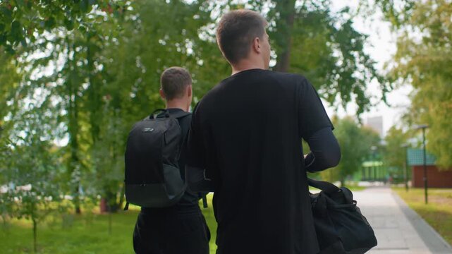 two white men walking together down treelined park path with backpack and duffel, relaxed conversation and casual pace, lamp posts and benches frame scene, summer light, scenes suggest students