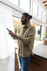 Indian man holding tablet standing by kitchen island with potted plant, photography light stand