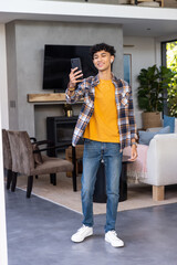 Naklejka premium Asian male student standing in living room holding smartphone while wearing wireless earbuds