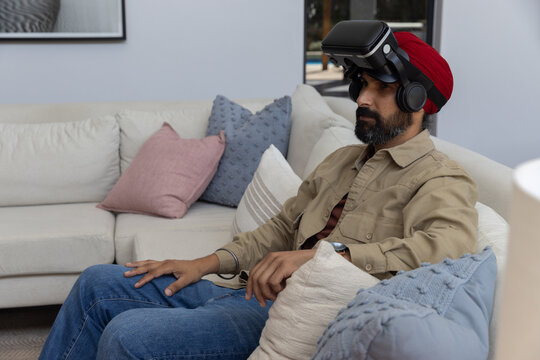 Indian man wearing red turban using virtual reality headset sitting on sofa, copy space - Powered by Adobe
