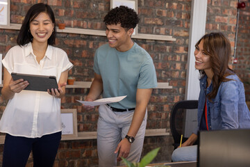 Diverse coworkers collaborating on project at desk in office with tablet, papers, pencil, laptop