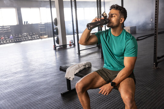 Asian man holding shaker bottle and drinking water while sitting on bench at gym, copy space