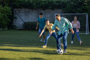 Diverse family playing soccer on grass field next to brick wall with soccer ball and goal net