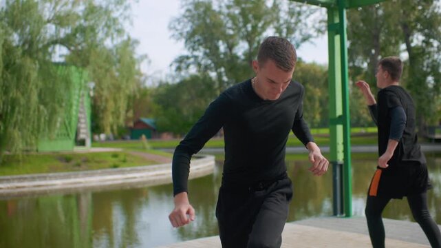Young white man dancing by lake, solo practice focused on movement and expression at green pavilion, quiet training in black outfit and sneakers, reflective mood with trees and calm water background