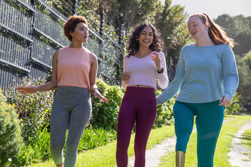 Diverse female friends walking along park trail by metal fence wearing sportswear, smartwatch