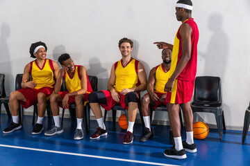 Diverse male basketball teammates sitting on sideline in gym laughing, pointing at basketballs