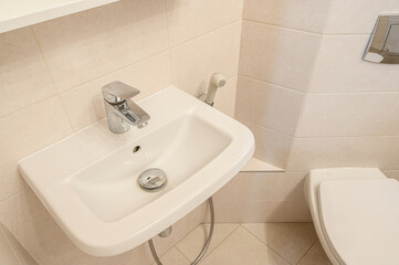 Wall-mounted white ceramic sink with chrome faucet; adjacent to toilet with concealed cistern; beige tiled walls and floor; includes hygienic shower attachment.
