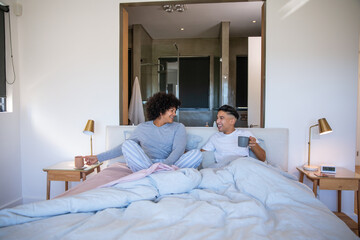 Fototapeta premium Male couple sitting in bed holding mugs, smiling and chatting by wooden nightstands with gold lamps