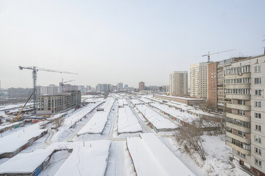 Winter urban landscape: snow-covered rooftops, residential buildings, construction cranes, undeveloped land plots, distant skyline, overcast sky, bare trees lining the area.