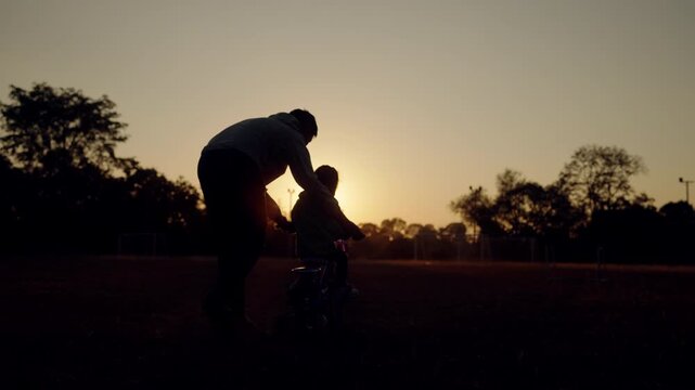 Silhouette of a father helping daughter ride a bike during sunset with sunlight shining through. Powerful image of parenting, support, and growth.