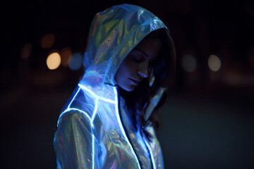 Young woman wearing a futuristic holographic iridescent raincoat illuminated by vibrant blue led lighting stripes while standing outdoors in the dark city environment