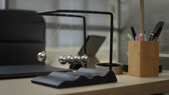 Closeup no people shot of Newtons cradle with five metallic spheres in motion at office desk, demonstrating physics principle of conservation of energy
