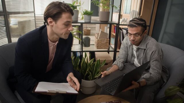 Gen Z African American man as junior specialist sitting in front of laptop in modern coworking space and asking his young Caucasian supervisor browsing through business documents to help him with task