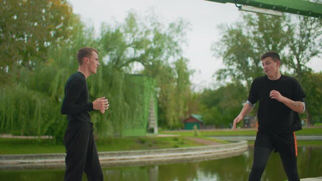 caucasian men sparring by pond, intense kick and punch exchanges under green pavilion, autumn trees and calm lake backdrop, trainer demonstrating technique, student evading, sparring partners trading