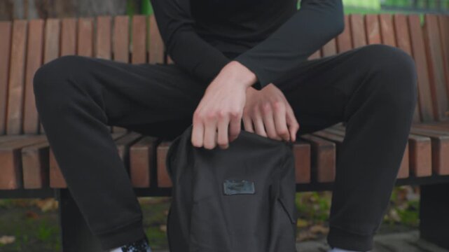 man seated on bench pulls helmet student commuter pauses with black backpack, closeup hands unzip bag, retrieves protective helmet, adjusts strap, casual hoodie and sneakers, autumn park atmosphere,