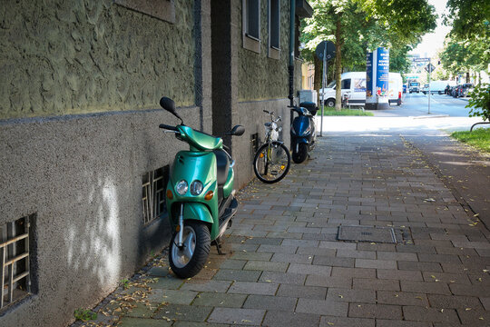 Ein gr&uuml;nfarbener Motorroller der Marke Yamaha Modell Neos an einer Hauswand vor einem Fahrrad und einem Motorroller der Marke Piaggio Modell ZIP S 50