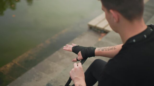 white male securing hand wraps near lake, tattooed forearms, methodical fingers, black wrap on concrete steps underground fighter prepping, coach inspection, competitor routine, ritual taping,