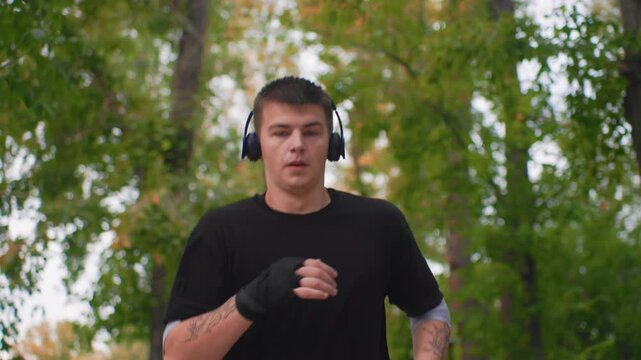 Caucasian man shadowboxing with headphones outdoors, hand wraps visible, focused facial expression, leafy park backdrop, punch combinations timed to music, energetic training session, autumn tones,