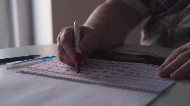 Man In Casual Shirt Plays Bingo Game, Man Sitting At Kitchen Table Marks Bingo Card With Marker, Casual Man Wearing Shirt Carefully Marks Numbers On Bingo Card During Game Night
