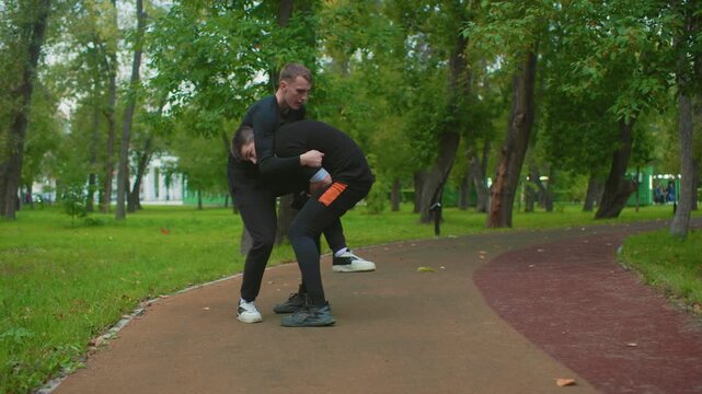 white male fighters grappling on tree lined path during aggressive sparring, clinch escalates into takedown and ground control, partner applying hold while training coach observes, raw intensity