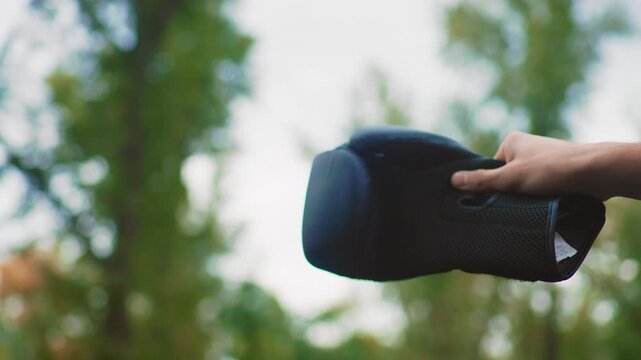 Closeup of handheld speaker amidst foliage, Black mesh speaker held by palm with blurred greenery behind, Outdoor scene displaying portable speaker gripped by hand under leafy trees in daylight