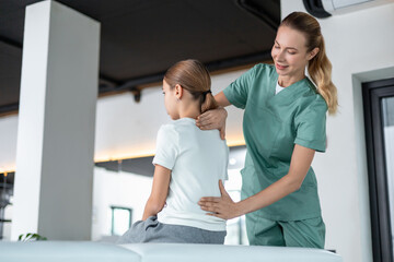 Fototapeta premium Woman physio in medical uniform checking girl spine in rehab room