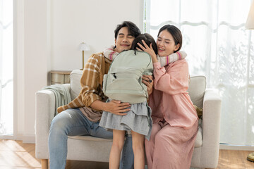 Happy Asian family hugging young daughter with school backpack on sofa in bright living room showing love and warm bonding with parents