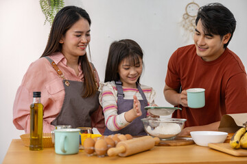 Happy Asian family cooking together in kitchen at home with warm smile and joyful expression while sifting white flour into bowl for baking cake or bread with fresh ingredient on wooden table