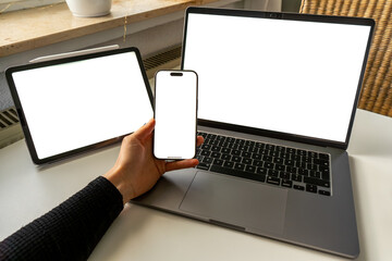 POV minimal workspace mockup with laptop, smartphone, and tablet screens blank. Hand holding tablet, modern flat lay for digital UI showcase.