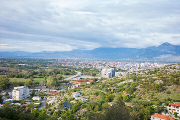 Obraz premium Wide panoramic view of a city nestled between lush green hills and expansive valleys, with residential neighborhoods, winding roads, and distant mountains under a dramatic cloudy sky
