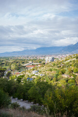 Fototapeta premium Wide panoramic view of a city nestled in a valley, seen from lush green hills overlooking urban buildings, winding roads, and distant mountains under a dramatic cloudy sky