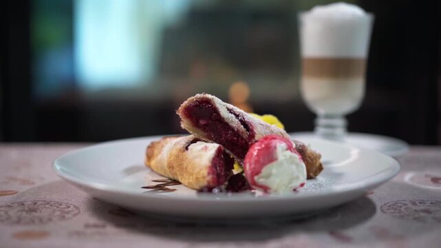 Close-up of a delicious berry strudel on a white plate with a scoop of vanilla ice cream, berry sauce, and powdered sugar. A glass of latte coffee is visible in the background