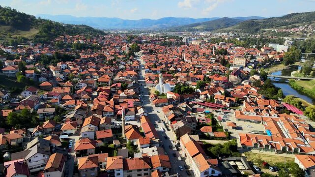Drone view over the city center of Visoko, showing dense red roof houses, main streets, mosques, and surrounding hills, highlighting the urban layout within a mountainous landscape.