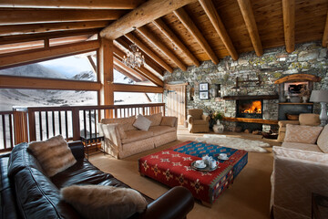 Naklejka premium Luxury ski chalet interior with wide views over snow covered mountain. Comfy sofas and blazing log fire with table set for afternoon tea. 