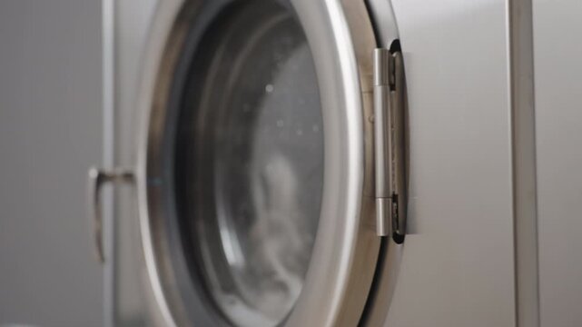 Close up right to left view of washing machine door showing stainless handles and glass, cloth tumbling inside with bubbling soap water, detailed components of industrial washer during wash cycle