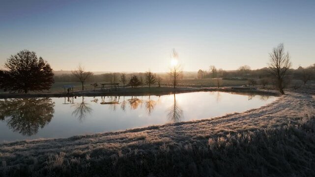 Aerial view of frosty morning landscape in Monpazier. Sun rises behind bare trees reflecting in calm pond waters. Winter scenery in Dordogne countryside with white hoarfrost covering the grass.