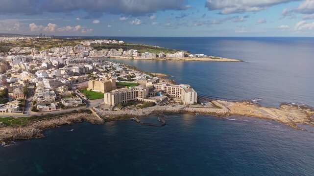 Orbital aerial view of Marsaskala, Malta, highlighting its picturesque coastal architecture, Mediterranean shoreline, and urban layout under a blue winter sky.
