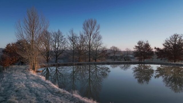 Cluster of leafless trees on icy grass bank near calm water. Drone shot capturing crisp winter atmosphere and frost texture on vegetation in Monpazier region.