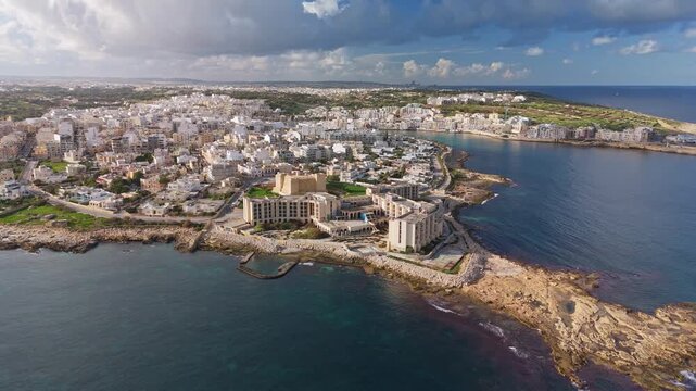 Orbital drone fly around Marsaskala, Malta, highlighting its coastal urban charm, Mediterranean architecture, scenic shoreline, and vibrant seaside landscape under a blue sky.