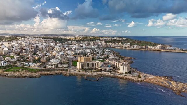 Orbital drone hyperlapse around Marsaskala, Malta, highlighting its coastal urban charm, Mediterranean waters, scenic peninsula, and vibrant harbor under a blue cloudy sky on a sunny winter day.