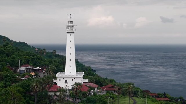 Aerial drone view of white lighthouse on tropical cliff overlooking the ocean, dramatic cloudy sky, coastal landscape in Indonesia.