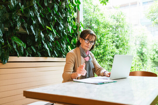 An older woman works on a laptop at a table in a bright, green indoor space with large plants in the background.